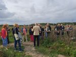 Een groep mensen staat op een pad in een open veld, luisterend naar een spreker die voor hen staat. De groep bestaat uit zowel mannen als vrouwen van verschillende leeftijden, gekleed in casual kleding. Op de achtergrond zijn bomen en een bewolkte lucht zichtbaar.