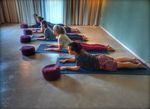 Een groep van vijf mensen ligt op blauwe yogamatten in een lichte ruimte, bezig met een yogapositie waarbij hun bovenlichaam omhoog is en hun armen gestrekt zijn. Voor hen liggen paarse yoga bolsters. Op de achtergrond zijn grote ramen met gordijnen van het Eemhuis zichtbaar.