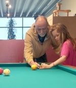 Een oudere persoon helpt een kind met het spelen van biljart. Ze staan samen aan de tafel, terwijl ze hun handen op de keu en de bal leggen.