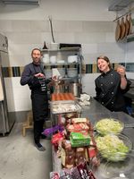 Twee enthousiaste koks in een keuken, bezig met het bereiden van maaltijden. De man in een schort houdt een pan vast, terwijl de vrouw naast hem vrolijk een drankje vasthoudt.