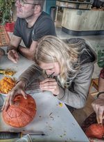Een jongen met lange blonde haren concentreert zich op het versieren van een pompoen, terwijl er een volwassene naast hem zit. Pompoenresten liggen verspreid op de tafel.