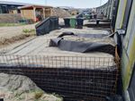 De grasheuvels voor onze huizen in aanbouw. Het terrein toont een uitgraving met een staalconstructies en zwarte doeken, dit alles gevuld met wit zand. Op de achtergrond zijn een houten fietsenstalling en afvalcontainers zichtbaar