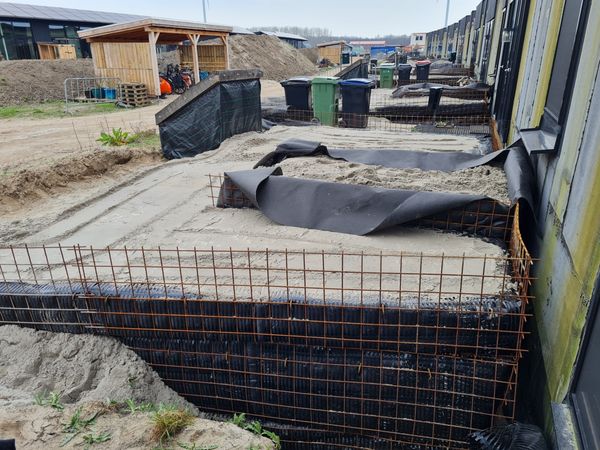De grasheuvels voor onze huizen in aanbouw. Het terrein toont een uitgraving met een staalconstructies en zwarte doeken, dit alles gevuld met wit zand. Op de achtergrond zijn een houten fietsenstalling en afvalcontainers zichtbaar