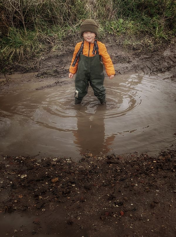 Een jongen in een oranje jas en groene laarzen staat vrolijk in een modderige plas. Hij kijkt lachend naar de camera, omringd door gras en struiken.