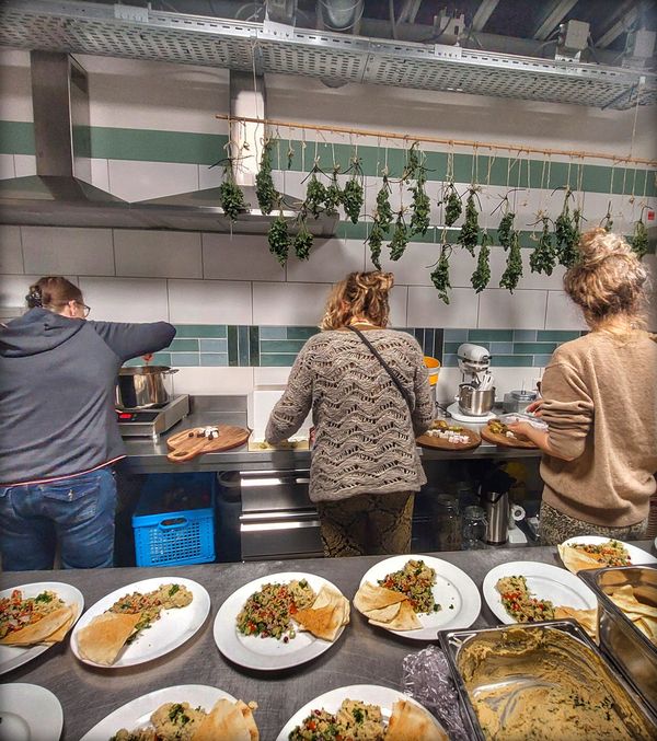 Drie mensen staan in de keuken, bezig met het bereiden van voedsel. Op de werktafel staan borden met salade en andere gerechten.