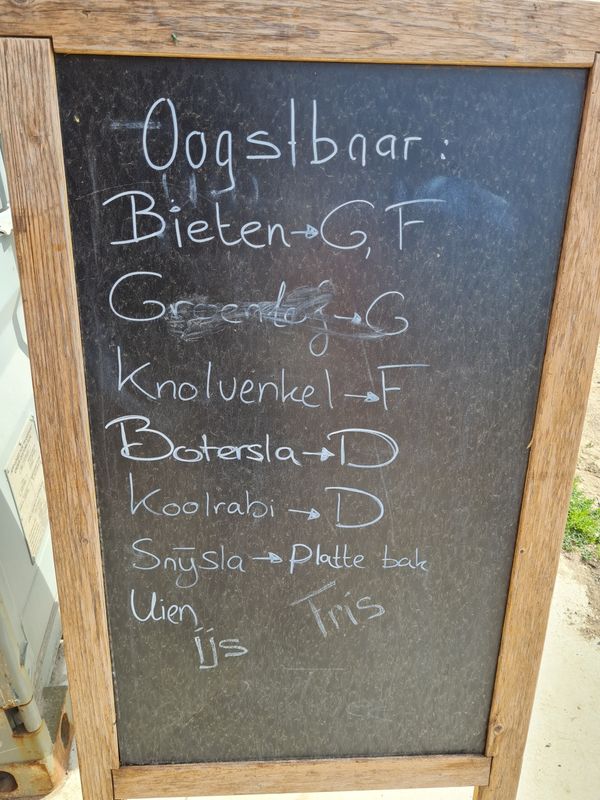 Een houten bord met een krijtbord oppervlak, waarop verschillende groenten en hun oogsttijd zijn geschreven, zoals bieten, knolvenkel en uien.