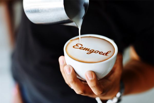 Een hand houdt een witte kop koffie met een latte art ontwerp waarop het woord 'Eemgoed' staat geschreven in chocoladekleurige melk.