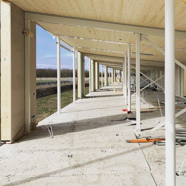 Een deels afgewerkt longhouse, met houten plafonds en metalen steunpilaren. De vloer is van beton en er zijn verschillende bouwmaterialen en gereedschappen zichtbaar.