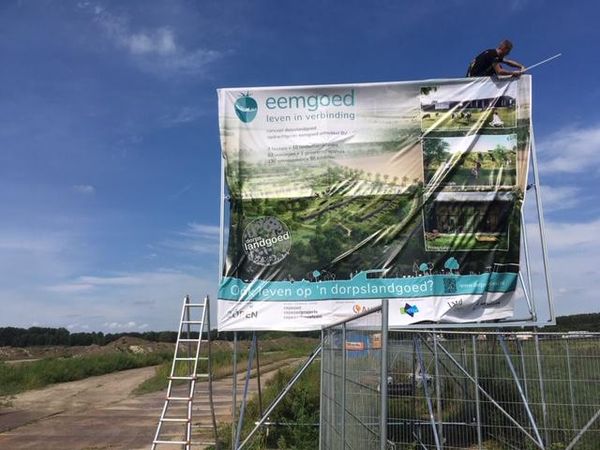 Een groot informatiebord met de tekst 'eemgoed leven in verbinding' en afbeeldingen van een groen landschap. Een persoon is bezig met het bevestigen van een doek aan het bord.